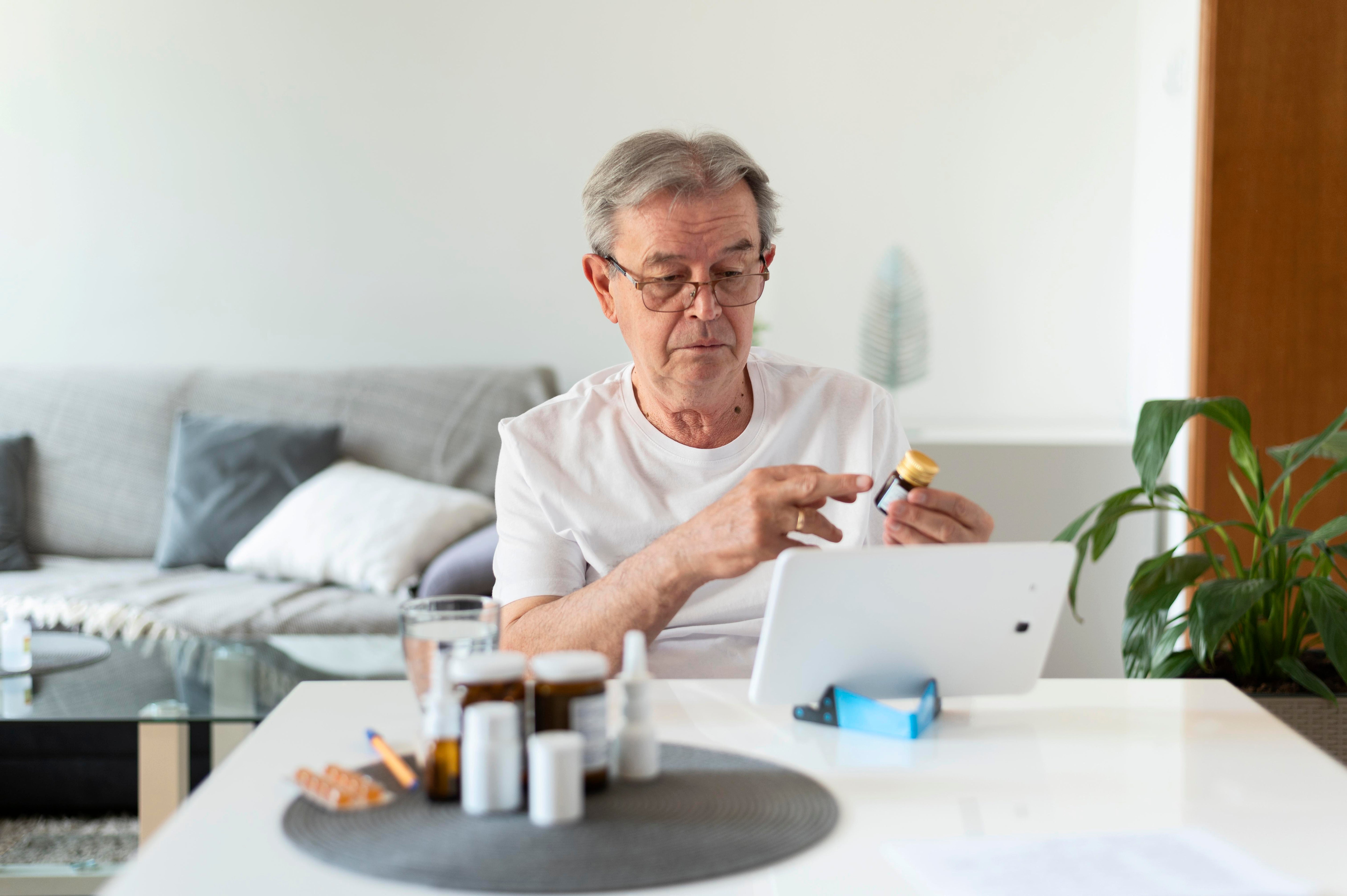 Man using a tablet at a table with various items including bottles and a plant in the background.